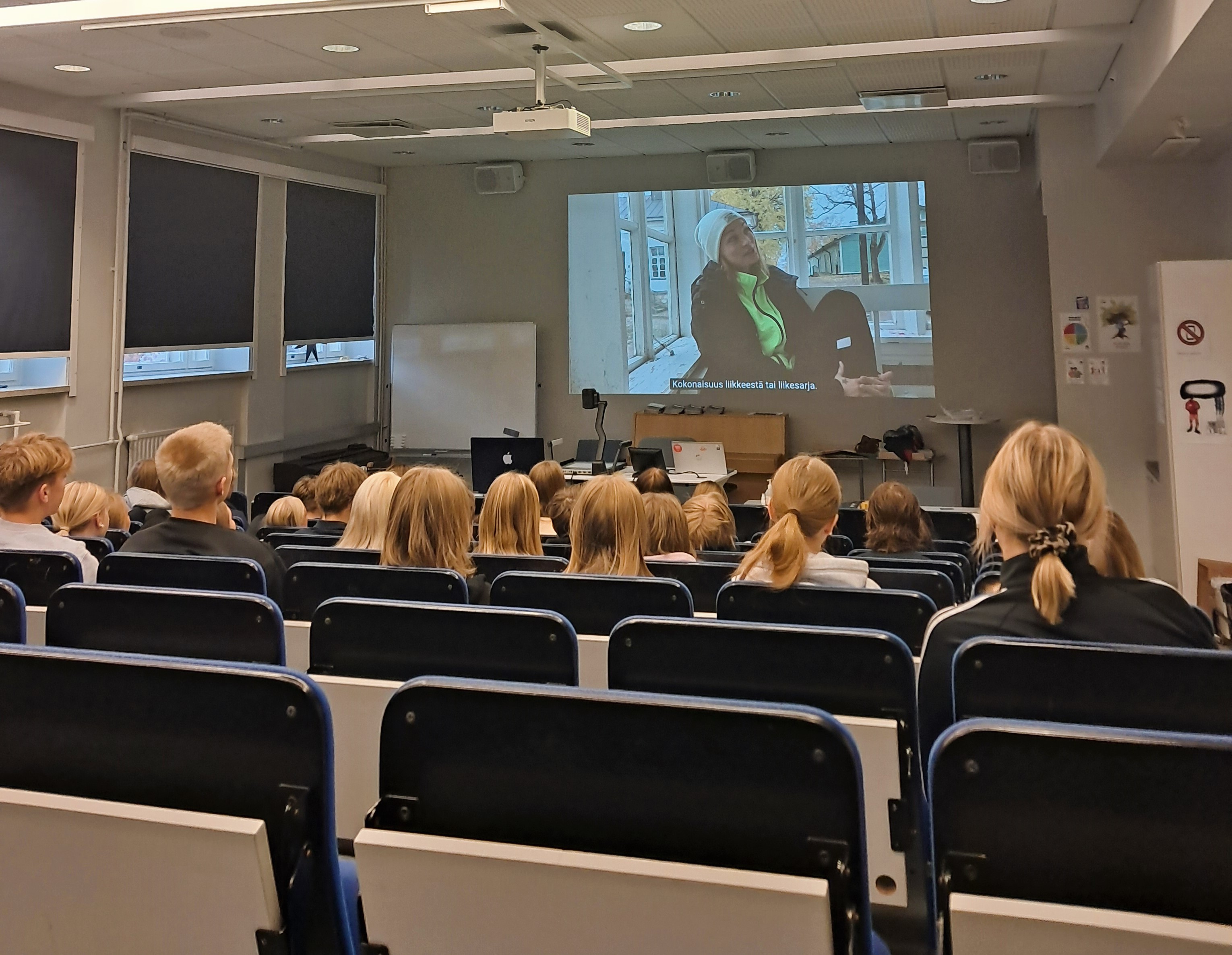 Oppilaita auditoriossa katsomassa kankaalle heijastettua videota.